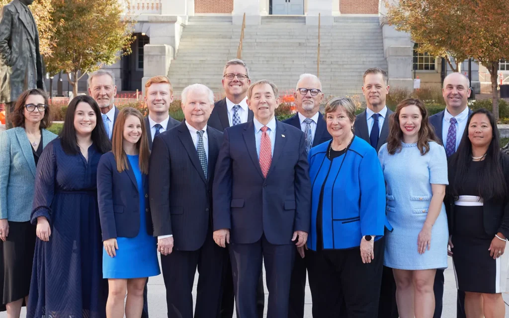 Group portrait of professionally dressed men and women standing outdoors in front of a building with stone steps and autumn trees.