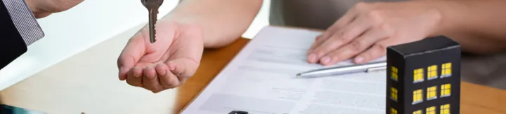 Person handing over a key to another individual over a contract with a small house model on the table.