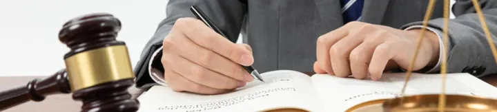Person in suit writing in open book, with gavel and scales of justice nearby.
