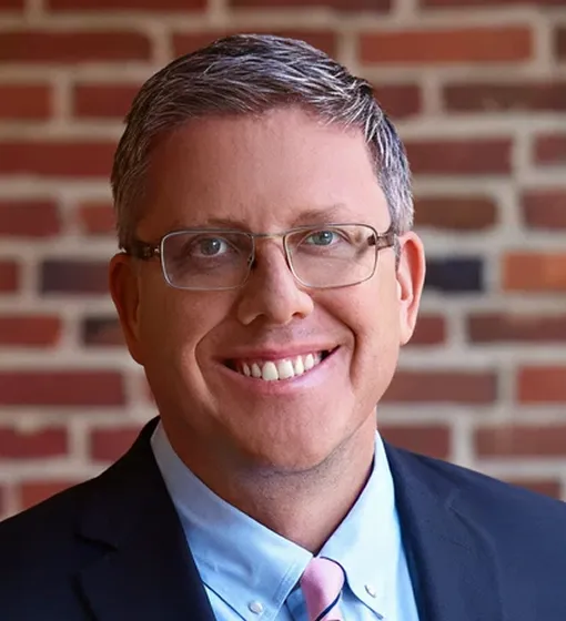 Professional portrait of a middle-aged man with glasses, wearing a suit and tie, smiling against a brick wall background.