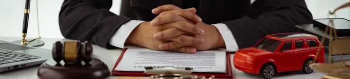 Person in dark suit with clasped hands behind legal documents, gavel, toy car, and scales of justice.
