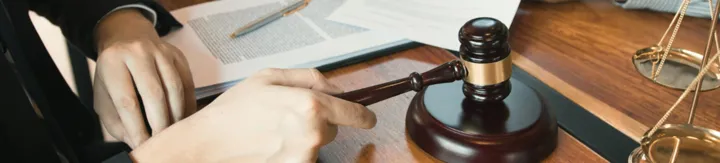 Person holding wooden judge's gavel over sound block on desk with legal documents.