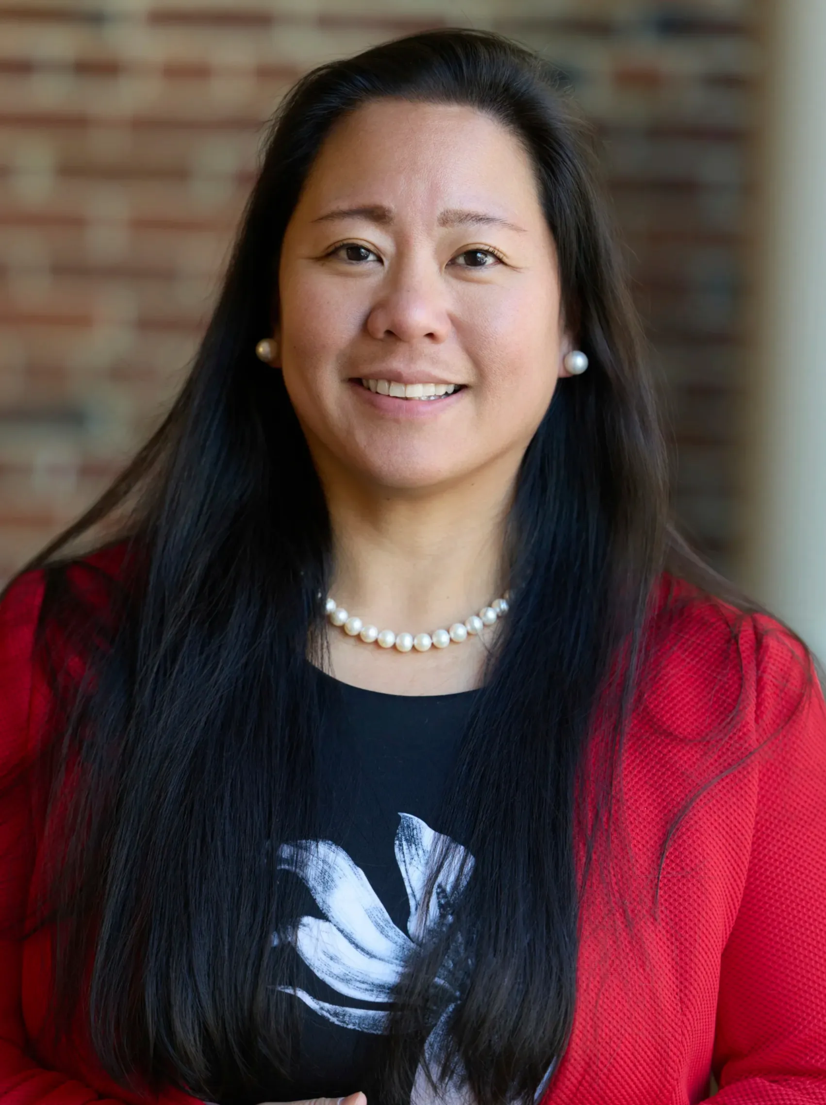 Professional portrait of a smiling woman with long black hair, wearing pearl earrings, necklace, red blazer, and black top.
