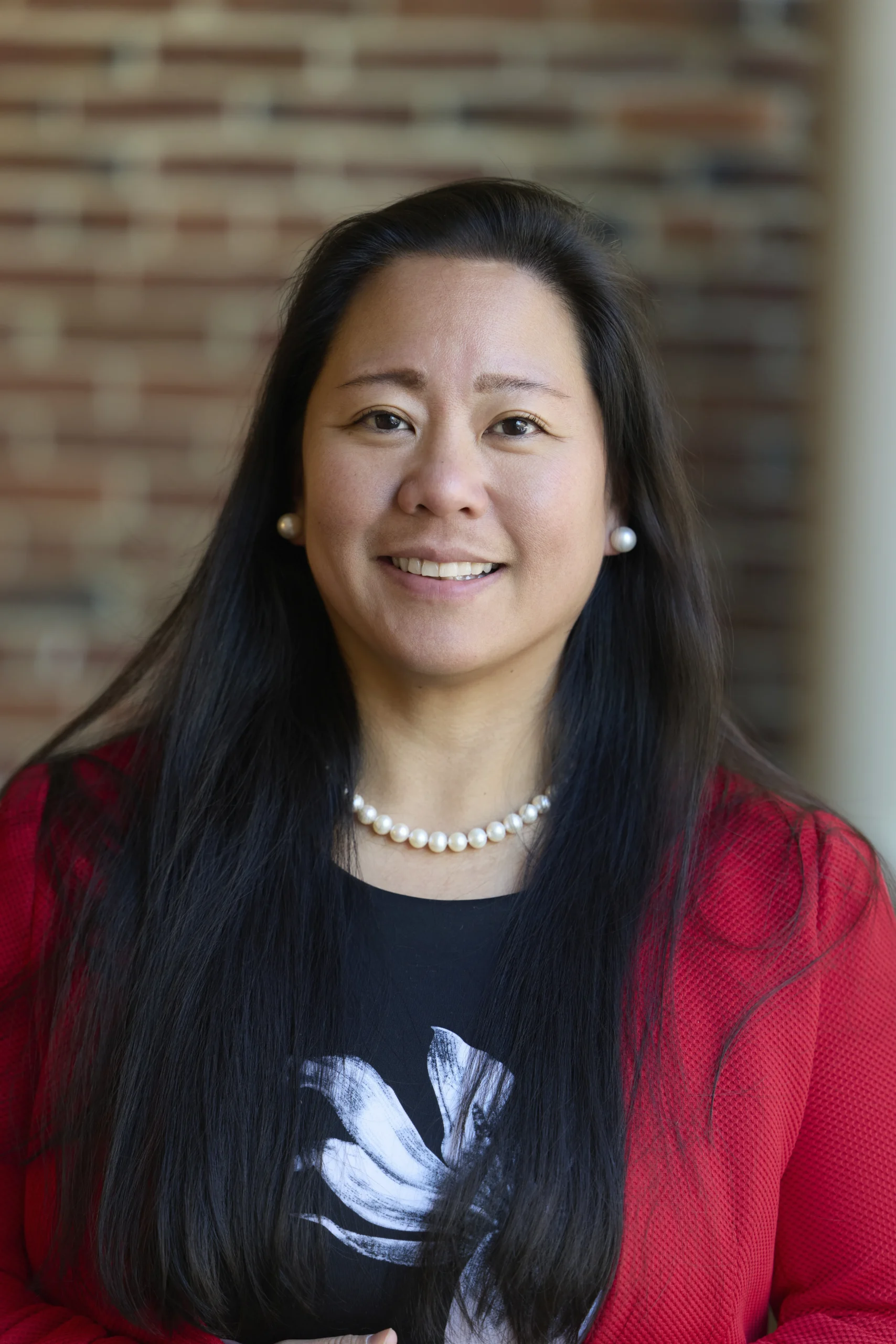 Professional headshot of a woman with long black hair, wearing a red blazer, pearl necklace, and earrings, smiling against a blurred brick wall background.
