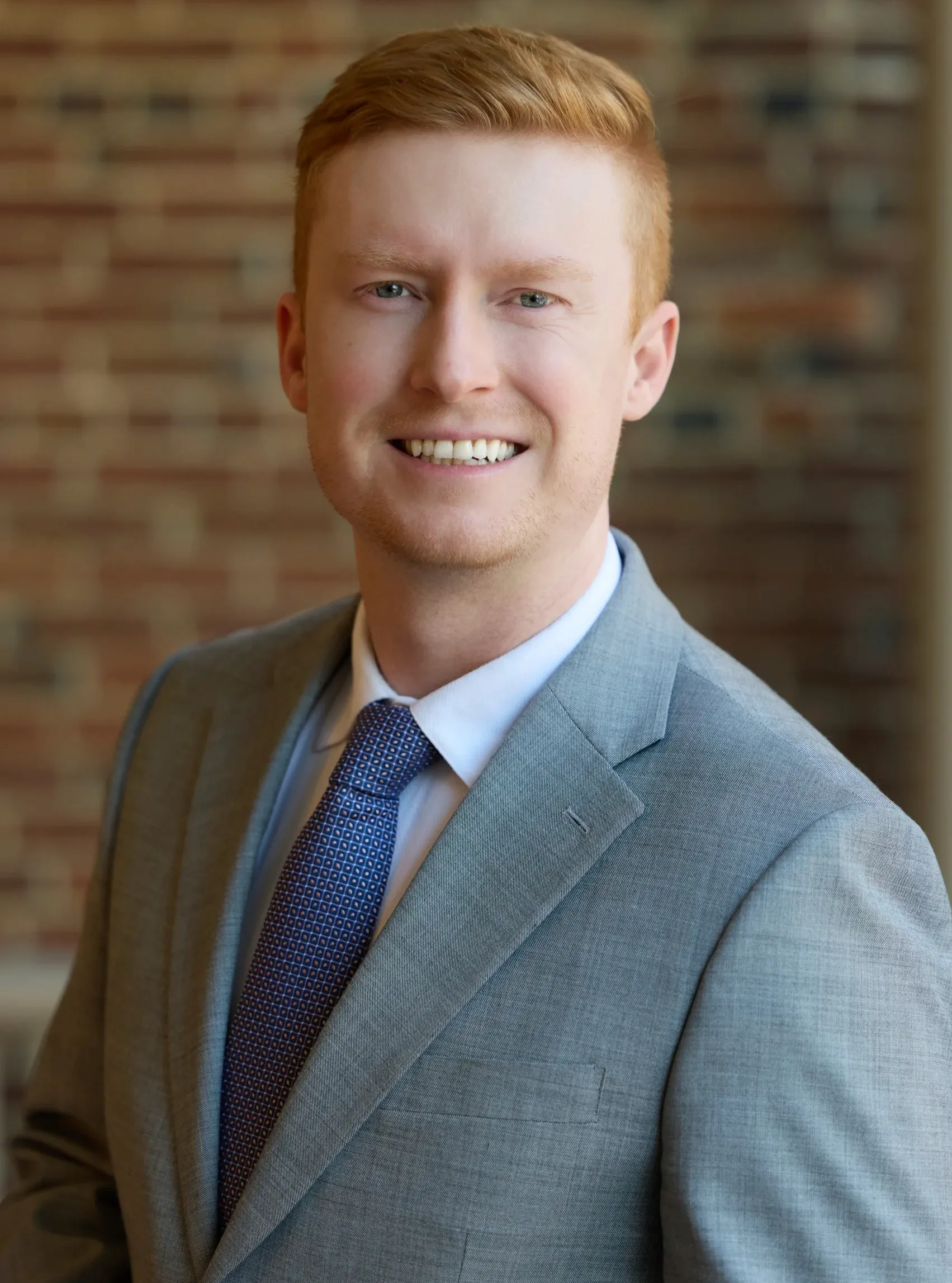 Professional headshot of a smiling red-haired man in a gray suit and patterned tie.