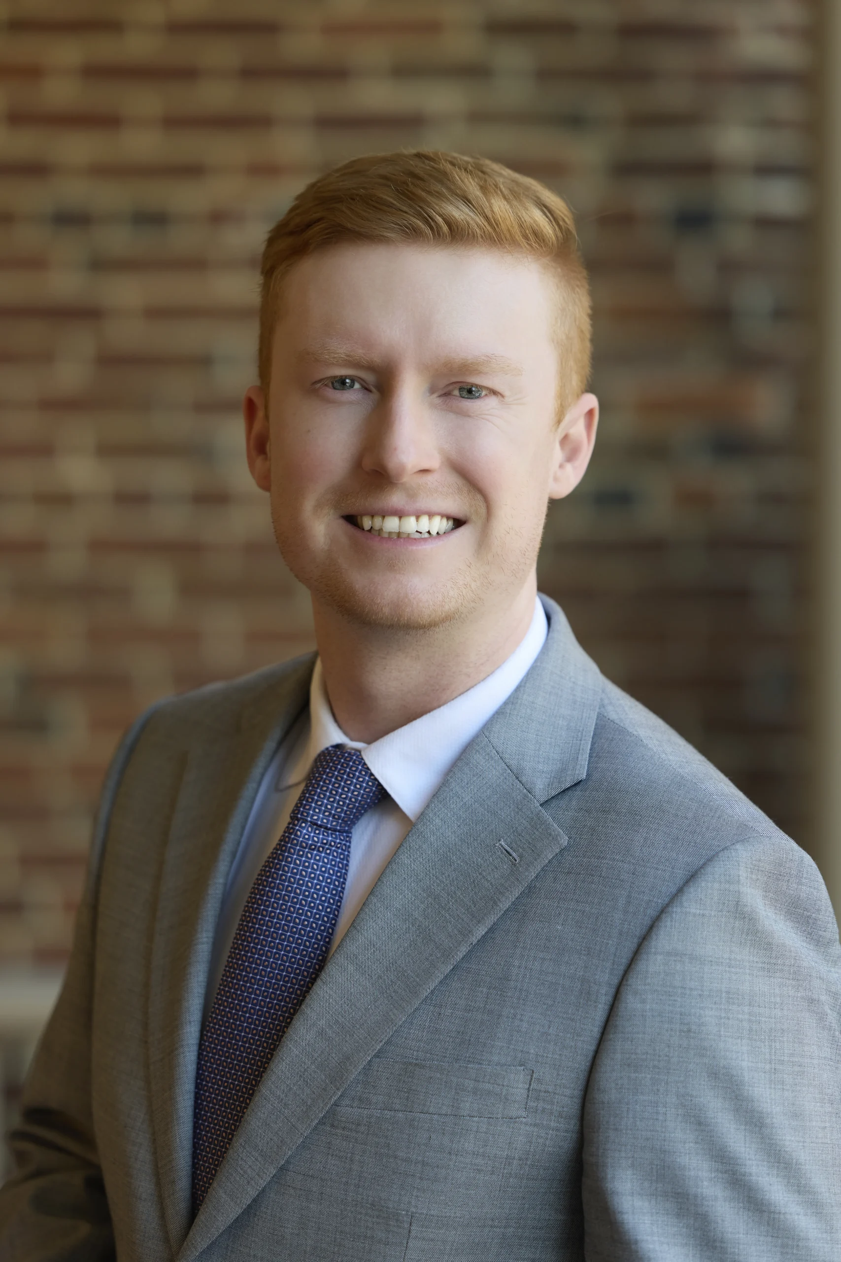 Professional portrait of a smiling red-haired man wearing a gray suit and blue patterned tie.