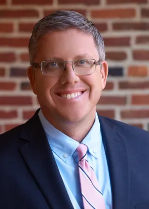 Professional headshot of a smiling man wearing glasses, a navy suit, light blue shirt, and pink striped tie against a brick wall background.