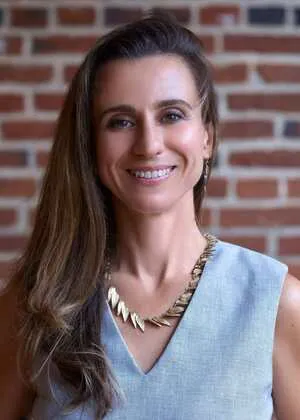 Portrait of a smiling woman with long brown hair, wearing a light blue sleeveless top and gold necklace, standing against a brick wall background.