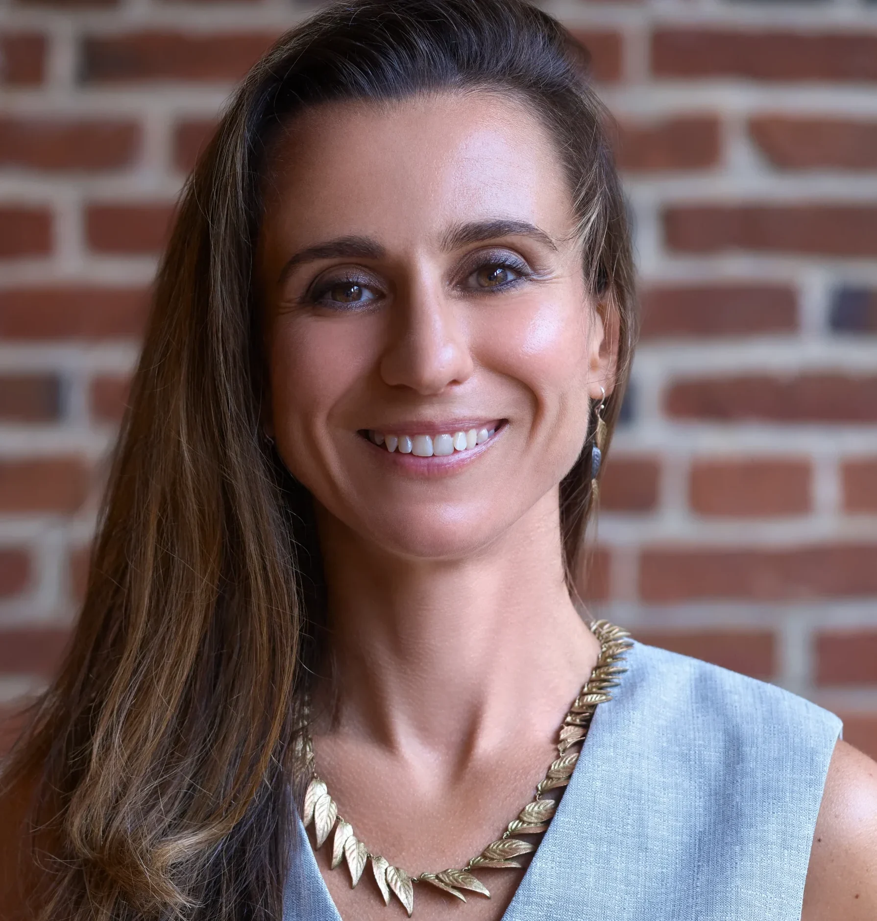 Professional headshot of a smiling woman with long brown hair, wearing a gray sleeveless top and gold leaf necklace.