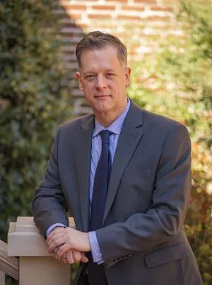 Professional middle-aged man in a suit and tie, standing outdoors by a brick wall.