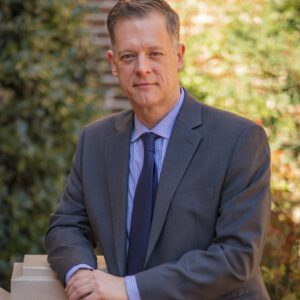 Middle-aged man in a gray suit and blue tie standing outdoors with blurred greenery background.