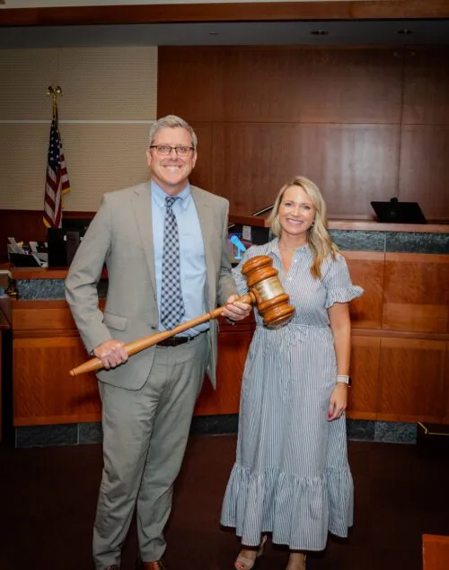 Two professionally dressed individuals standing in a courtroom, one holding a large wooden gavel.