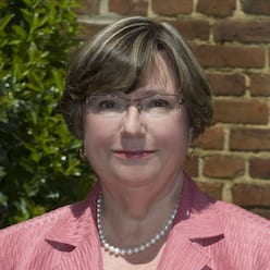 Portrait of a woman with short hair, glasses, pink blazer, and pearl necklace outdoors.