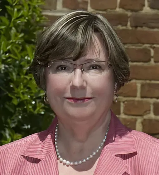 Professional portrait of a middle-aged woman with short hair, glasses, pearl necklace, and pink blazer.