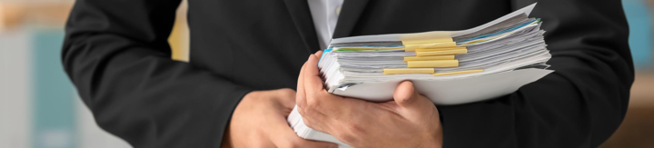Person in black suit holding a large stack of clipped documents and papers.