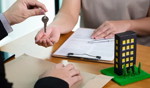 Two people exchanging a key over a table with documents and a miniature building model.