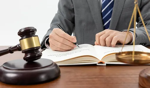 Person in gray suit writing in an open book with a wooden gavel and brass scales on desk.