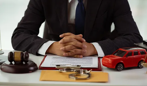 Person in dark suit sitting at desk with legal documents, gavel, handcuffs, and toy car.