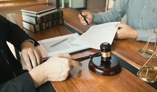 Two individuals at a wooden desk with legal documents, a gavel, law books, and scales of justice.