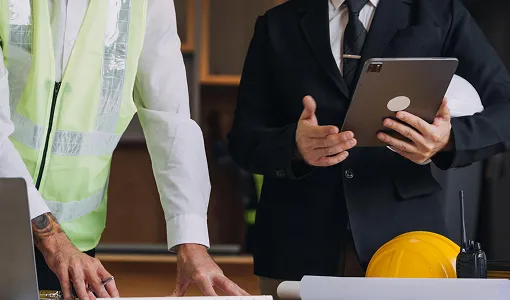Two professionals, one in a reflective safety vest and the other in a black suit, reviewing plans with a tablet and construction helmet on the table.