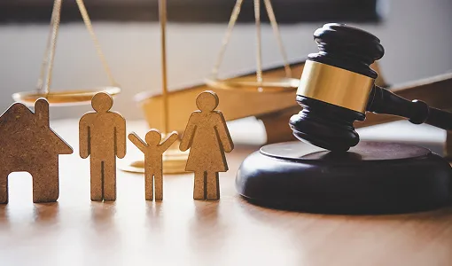 Wooden family figures and house model beside a judge’s gavel and golden scales of justice.