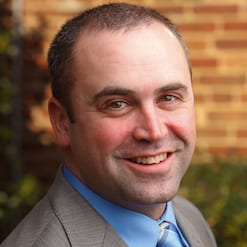 Headshot of a smiling man in a gray suit and blue tie with a brick wall background.