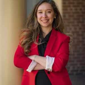 Professional young woman with long hair, wearing a red blazer and black blouse, smiling.