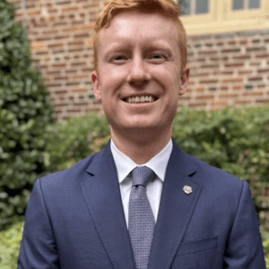 Portrait of a young man in a blue suit and tie, smiling outdoors near brick wall and greenery.