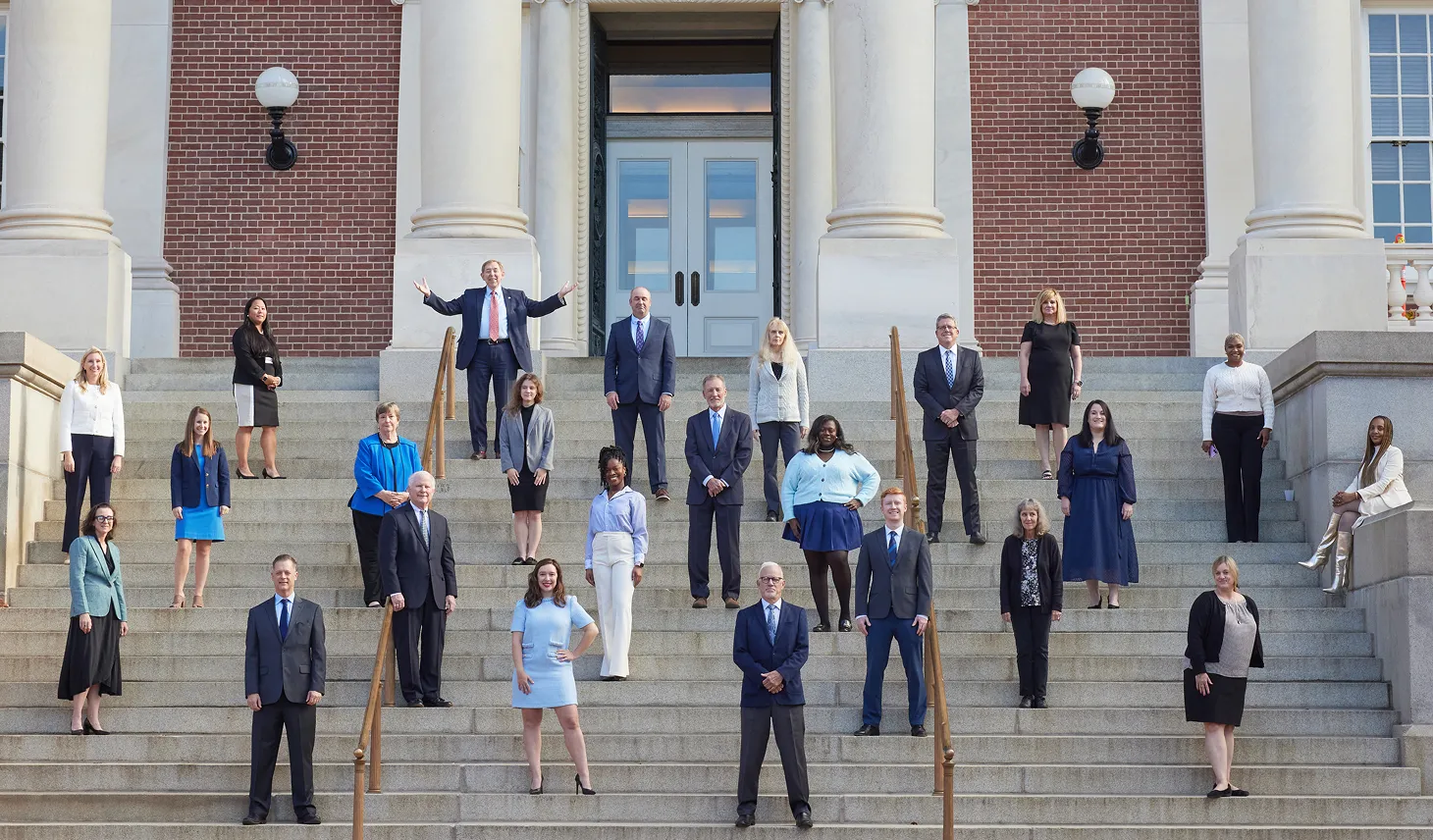 Group of professionally dressed men and women standing on wide stone steps in front of a brick building with large white columns and double doors.