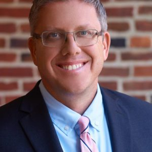 Professional headshot of a smiling man wearing glasses, a navy suit, light blue shirt, and pink striped tie, with a brick wall background.
