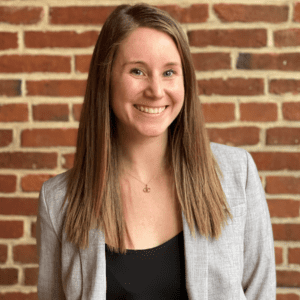 Portrait of a smiling woman with long brown hair wearing a gray blazer against a brick wall.