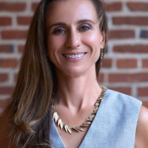 Portrait of a smiling woman with long brown hair, wearing a gray sleeveless top and gold necklace, standing against a brick wall.