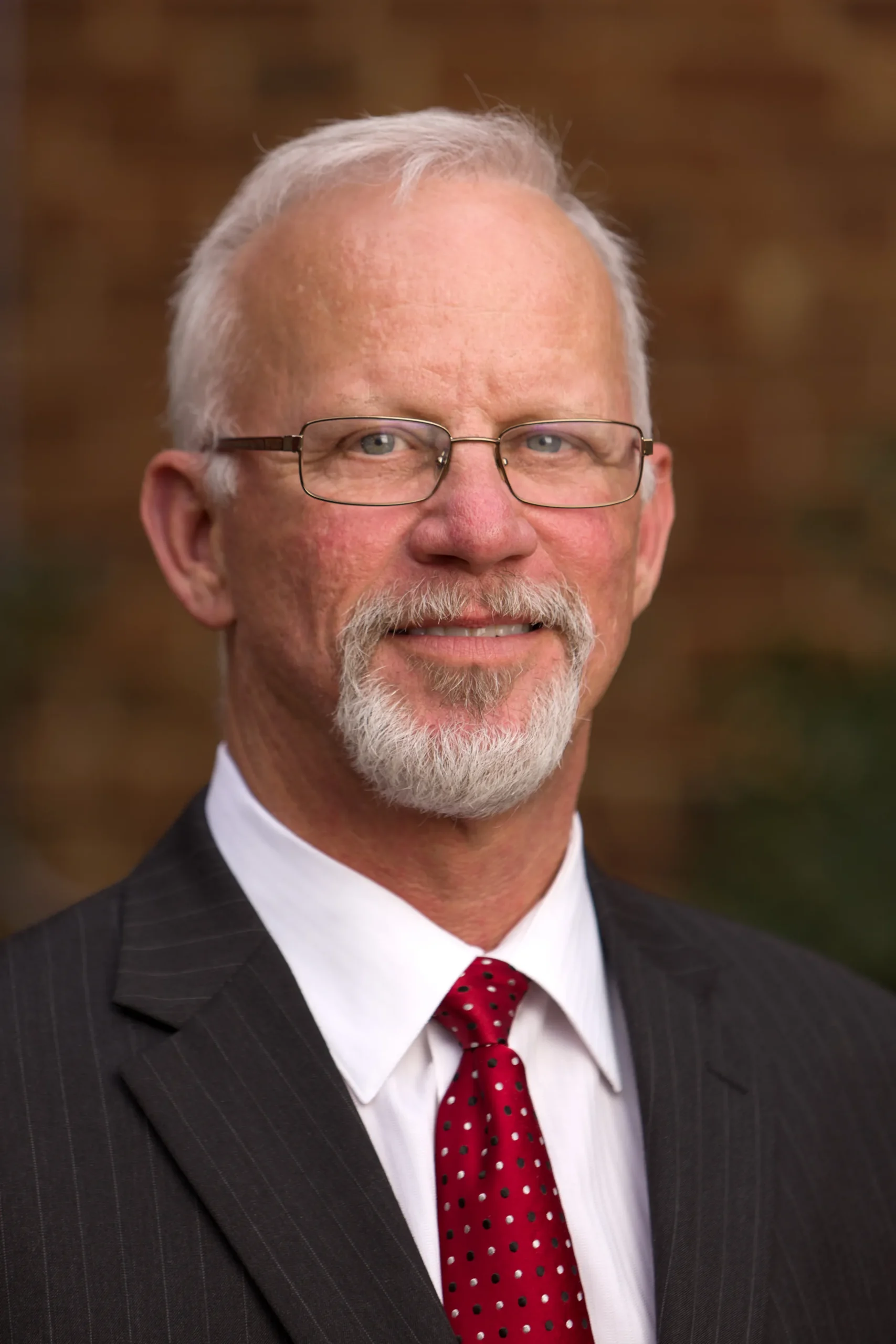 Portrait of an older man with white hair and beard, wearing glasses, a dark suit, white shirt, and red polka dot tie.