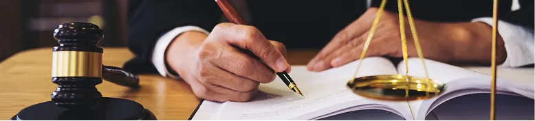 Person in formal attire writing in an open book with a gavel and golden scales on wooden desk.