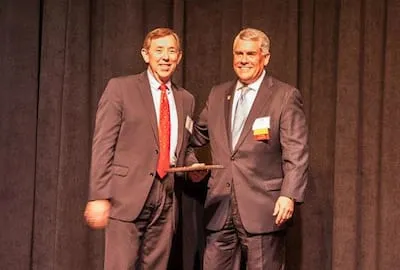 Two men in business suits stand on stage with a dark curtain backdrop, one holding a plaque.