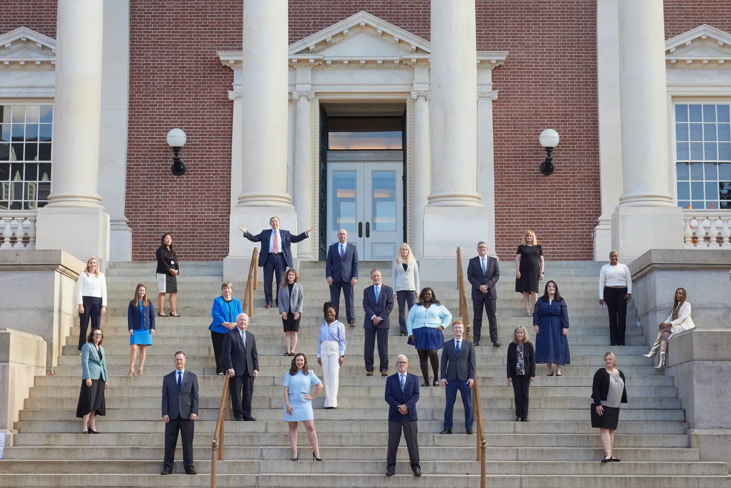 Group of professionally dressed individuals posing on large stone steps in front of a red brick building with white columns.