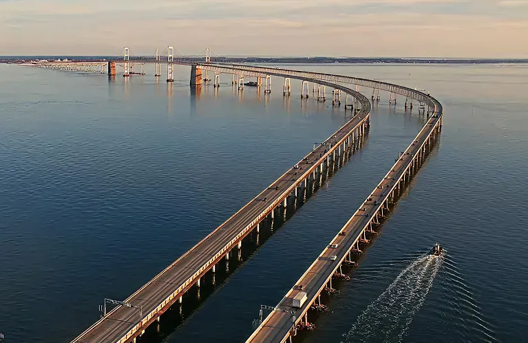 Aerial view of a long curved bridge over calm water with vehicles and a boat passing underneath.