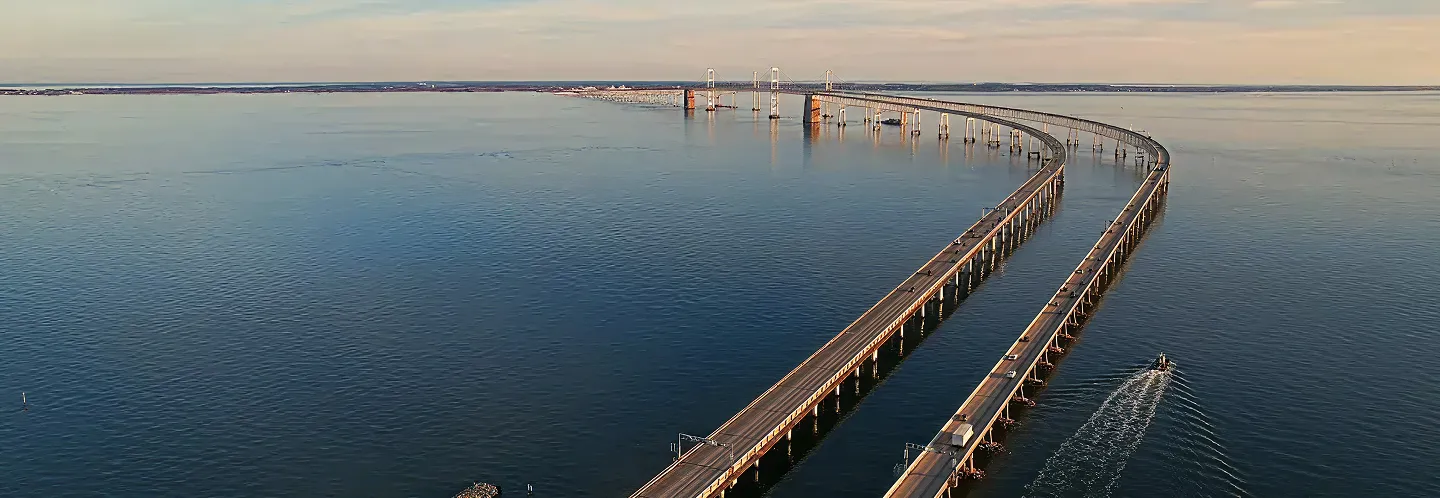 Aerial view of a long, curved bridge over calm blue water with a boat nearby.
