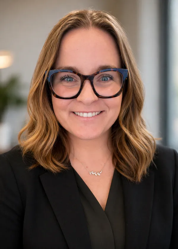 Professional woman with shoulder-length light brown hair, wearing black glasses and a black blazer, smiling.